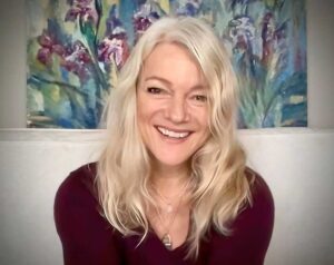 picture of woman with blonde hair and purple v neck jumper with a paining of flowers behind her